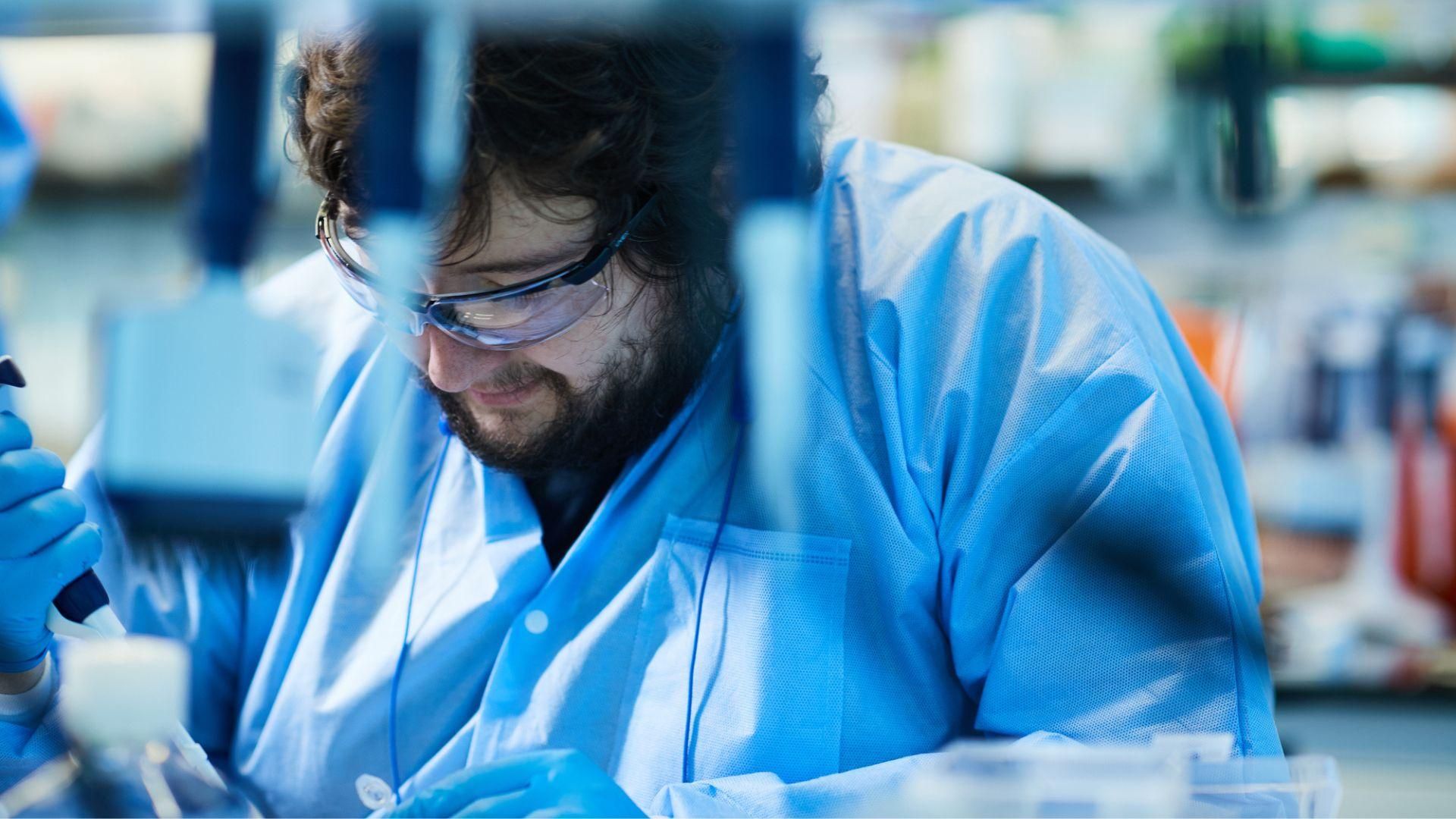 Nick Clark, with curly dark hair and beard, wearing blue gloves, lab outfit, and safety glasses, working with samples